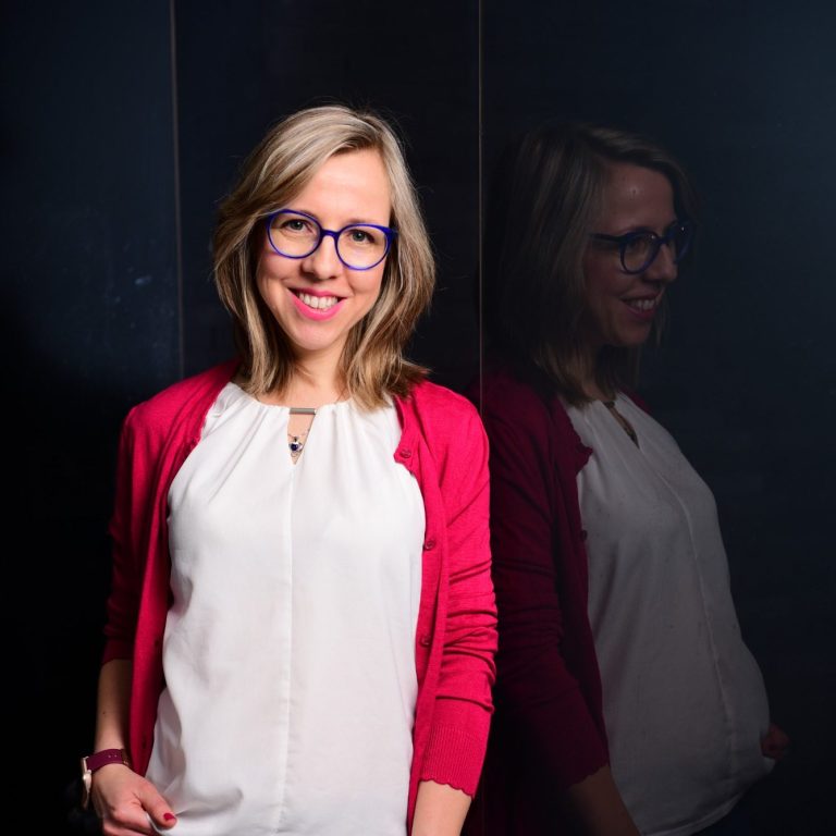 Eine lächelnde Frau mit blonden Haaren und Brille, trägt ein weißes Oberteil und eine rote Strickjacke.