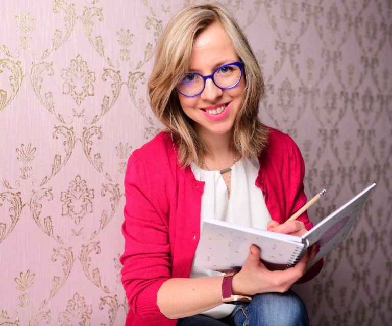Lächelnde Frau mit blauer Brille, hält ein Notizbuch und einen Stift, vor gemusterter Wand.