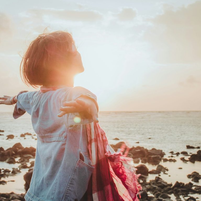 Frau mit offenen Armen am Strand, Sonnenuntergang im Hintergrund.