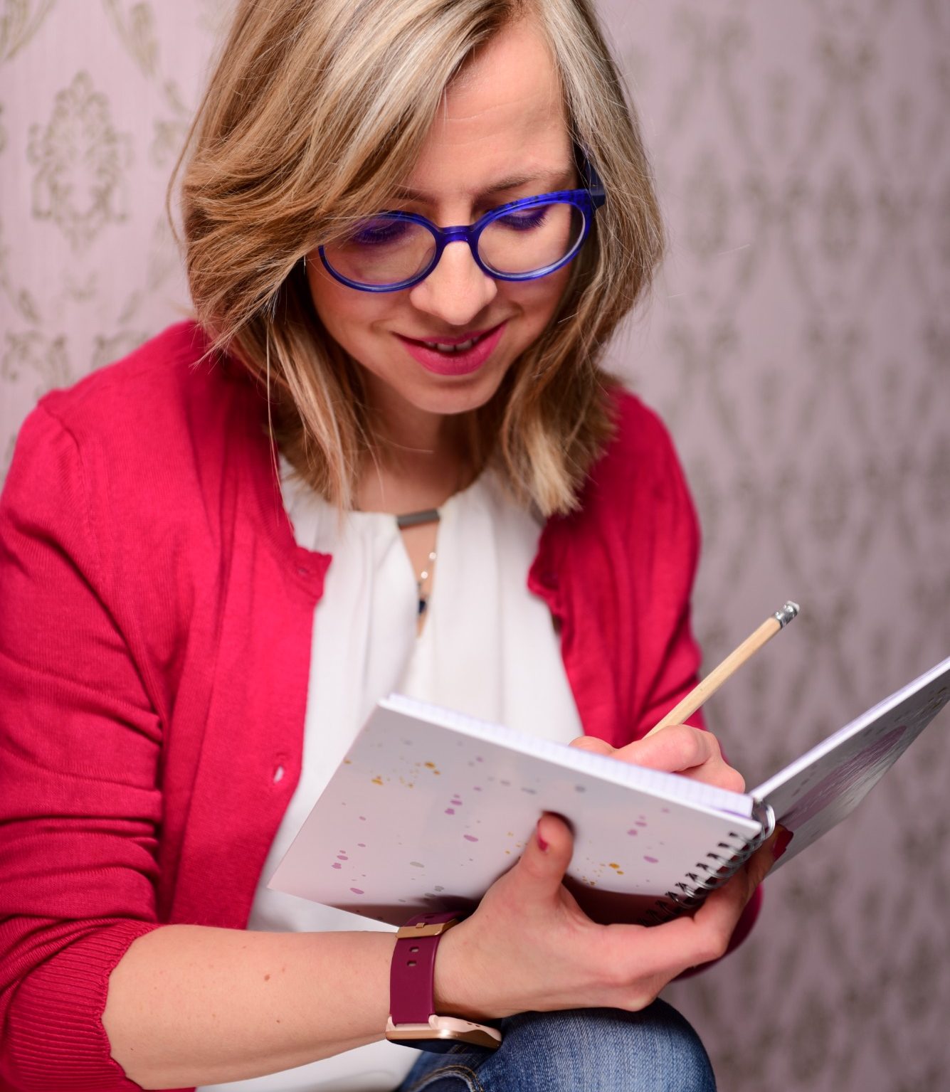 Junge Frau mit blonden Haaren, blauem Brillengestell und rotem Cardigan vor rosa Tapete.