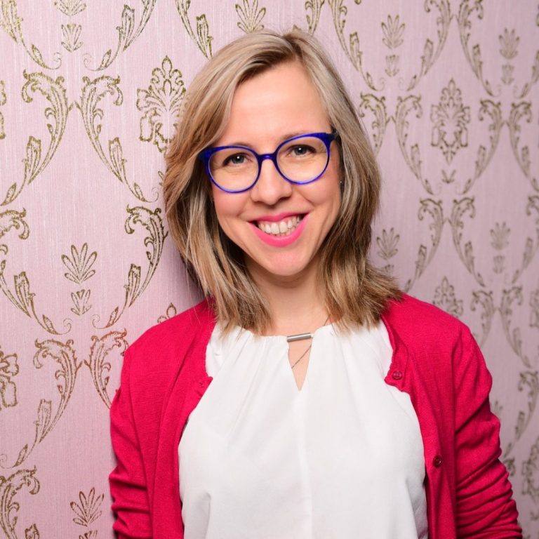 Frau mit blonden Haaren, blauer Brille und rotem Cardigan vor gemustertem Hintergrund.