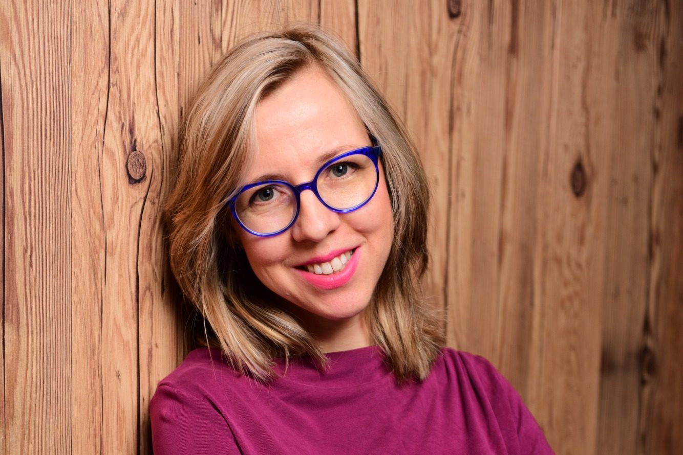 Junge Frau mit blonden Haaren und blauer Brille, lächelt vor Holzwand.