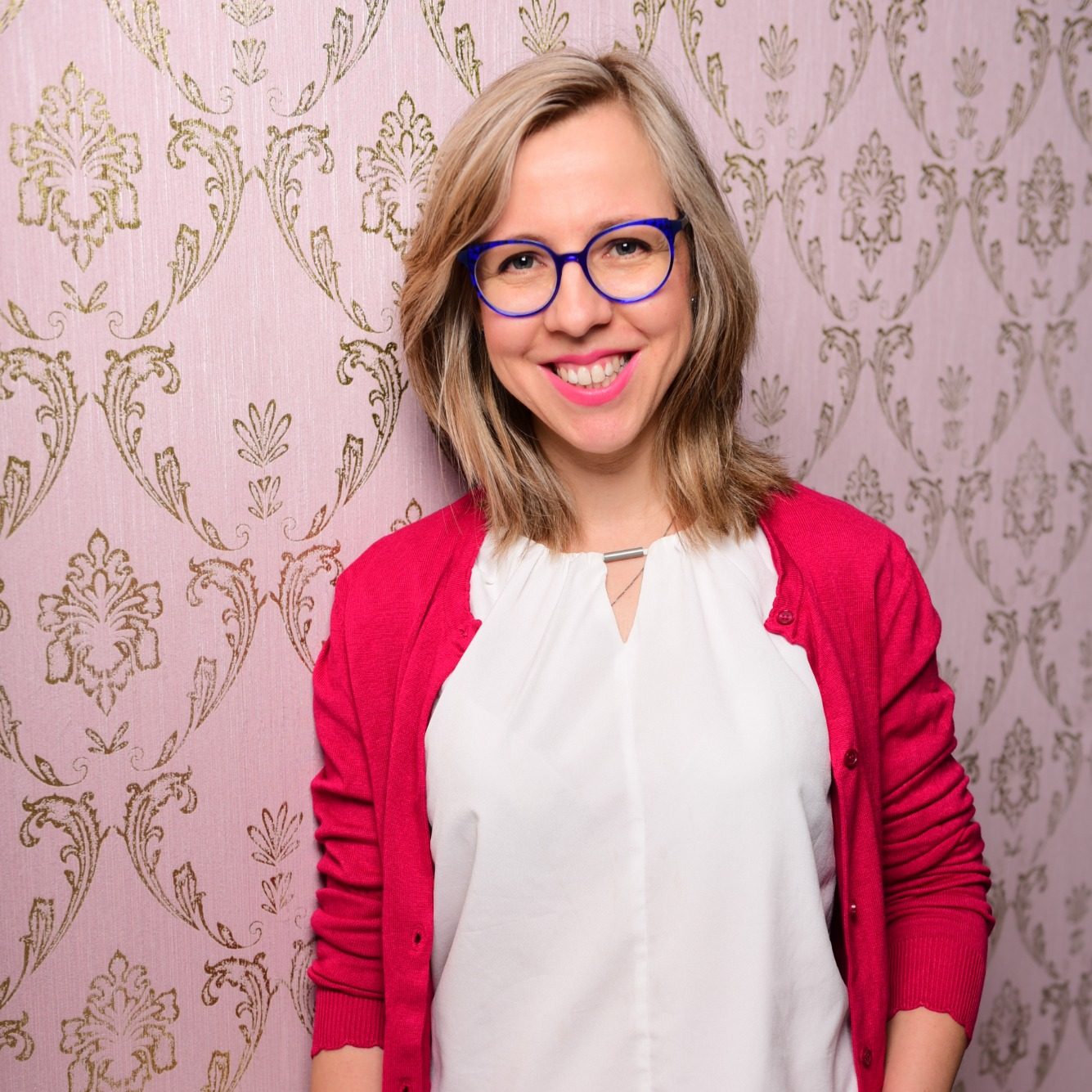 Junge Frau mit blonden Haaren, blauem Brillengestell und rotem Cardigan vor rosa Tapete.